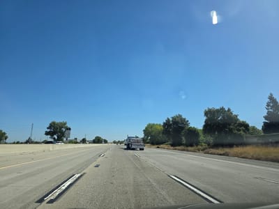 The media file shows a highway scene in Sacramento, California, under a clear blue sky, indicating daytime. The perspective is from inside a vehicle, suggesting the shot was taken while driving. The road is paved, with multiple lanes visible. On the left side of the road, there's a concrete barrier and beyond that, some greenery and power lines.
Further down the road on the left, a white sign is visible. Zooming in on the sign reveals text that appears to be regulatory, possibly related to vehicle restrictions or speed limits, although the details are difficult to discern. A truck is seen in the right lane of the highway ahead, carrying a load of what appear to be stacked materials, possibly building supplies. The truck is a semi-trailer truck.
To the right of the highway, there is a grassy area with dry brush, bordered by trees and a low wall. The overall impression is a typical highway commute in a semi-arid or warm climate, given the dry vegetation and clear skies. There are no people clearly visible in the frame, nor any explicit indication of a specific event. The scene conveys a sense of travel and the open road. There are some lens flares visible in the sky, indicating the sun is bright.