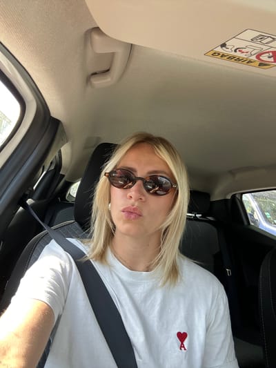A woman is seated inside a car, wearing sunglasses and a white t-shirt with a red heart logo, and a seatbelt. The interior of the car is visible, including the headrest, sun visor with an airbag warning label, and window. While it's impossible to confirm the exact location in Mougins, France, without more information, the picture suggests the woman is a passenger in a vehicle, possibly in transit.