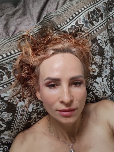 A woman with wet, reddish-blonde hair lies on her back on a patterned blanket. She is looking directly at the camera. Her makeup includes pink eyeshadow and lipstick. A silver necklace with a pendant is visible around her neck. The blanket has a brown and cream geometric pattern. The image is taken from a close-up perspective, focusing on the woman's face and upper body. There is no indication of the city of Kvartsitny, Russia, in the image.