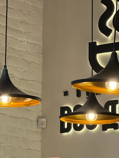 In the city of Косой Карьер Снт, Russia, an indoor scene is depicted. The focus is on several black pendant lights with gold interiors, hanging against a white brick wall on the left and a light gray wall on the right. The lights are illuminated, casting a warm glow. On the gray wall, there is a backlit black sign with stylized lettering, partially visible. A small white motion sensor is mounted on the brick wall. The overall impression is of a modern interior, possibly a restaurant or a commercial space, illuminated by ambient and task lighting.