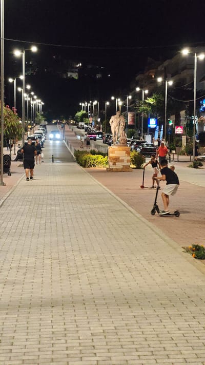 The image depicts a nighttime scene on a paved street lined with streetlights, a pedestrian walkway, and buildings. In the foreground, a person in a black t-shirt and shorts walks away from the camera on the illuminated walkway. Further down the street, two children are riding on electric scooters on the street. One child, in a black top and white shorts, is in the foreground on the right, and another child in a dark top and white shorts is further back on the left.
Several cars are parked along the street and some are in motion, with headlights visible. A prominent statue of two figures stands on a stone pedestal on the right side of the street, near some greenery. The atmosphere appears lively, with people out and about on a warm evening. The streetlights cast a bright glow, illuminating the scene against the dark night sky. The architecture of the buildings suggests a Mediterranean or European location. Some signs are visible, including a red stop sign and illuminated advertisements, but the text is not clearly legible. The overall scene captures a moment of urban nighttime activity.