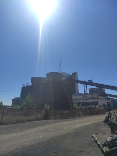 The image shows an industrial facility under a clear blue sky with bright sunlight. In the foreground, a dirt road leads towards the complex. A fence runs along the left side of the road, bordering an area with overgrown vegetation.
The main structure in the center appears to be a large industrial building with two prominent cylindrical cooling towers on top. A metal walkway or conveyor belt system extends from this building to another structure on the right. A crane is visible in the background. The buildings have a weathered, industrial appearance, with various pipes and support structures visible. The overall impression is of an active or recently active industrial site.
This facility is located in the city of Kyzyl, Kazakhstan, an industrial area known for its mining and processing operations. The scene depicts a typical industrial landscape, characterized by large-scale infrastructure and a focus on production. The bright sunlight suggests it is daytime, likely during warmer months, given the clear sky and the presence of green vegetation. There are no people visible in the image.
