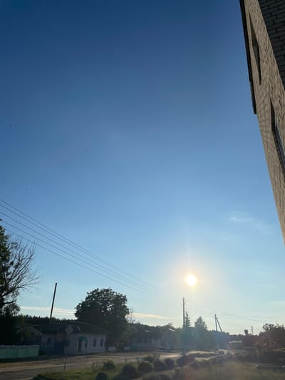 This is a vertical, eye-level, medium shot photograph taken outdoors on a clear, bright day. The scene depicts a rural area, possibly within the Азершчынскі сельскі Савет, Belarus, characterized by a clear blue sky with scattered, faint clouds near the horizon. The sun is visible as a bright, circular orb, slightly off-center to the right, casting a strong glare and illuminating the scene with natural light.
In the foreground, the edge of a brick building occupies the right side of the frame, its textured surface and diagonal line suggesting a building under construction or a partial view from a window. Below and to the left, a rural landscape unfolds. Several power lines stretch horizontally across the mid-ground, supported by utility poles. A single, bare tree with intricate branches stands on the far left.
A small cluster of low-rise buildings, appearing to be houses or village structures, are visible in the mid-ground, nestled amongst mature trees. A fence runs along the left side of the road, and some vegetation, including grass and bushes, is present in the foreground. The overall impression is one of a quiet, rural setting bathed in bright daylight. There are no people visible in the frame.