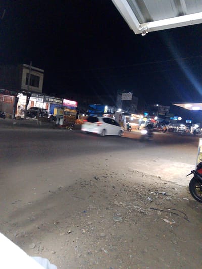 The image depicts a street scene at night in Gampongpaya, Indonesia. A white car is moving away from the camera on the right side of the road. On the left side of the road, there are several shops and food stalls. One stall is brightly lit with colorful umbrellas. A building with a prominent sign reading "WM BOUTIQUE" is visible. There are also several motorcycles and people scattered throughout the scene. The road appears to be paved with some debris on the side. The overall atmosphere is that of a busy commercial area at night.