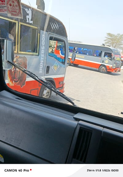 The image captures a street scene from the perspective of a vehicle's interior, looking out through its windshield. The foreground shows the dark gray dashboard of the vehicle, which has a textured surface, and a single air vent is visible on the right side. A windshield wiper rests horizontally across the lower part of the view through the glass.
Outside, two customized buses, commonly known as matatus, are visible. The bus on the left is closer and more prominent. It is predominantly gray with a red lower section. Its side is adorned with various graphics and text. Vertically aligned text reads "HUNTERS001" in white. A large, detailed graphic of a roaring lion's head is positioned on the lower front side of the bus. Above the windows, a white silhouette of a person aiming a rifle is displayed, and further up, a series of white claw marks are visible. Several stickers are affixed to the top-left of the bus's windshield, some partially visible: "DRIVE CAREFULLY", an "ISUZU" logo, information about "Breakdown / Accident" with a phone number "0743 1567" and "24 hrs", and "FASTEN YOUR BELT". The bus has gold-colored trim around its windows and a visible front wheel.
Further in the background, to the right of the first bus, another similar bus is seen. It shares a gray and red color scheme and features dark blue tinted windows. The ground beneath the buses appears to be light-colored asphalt or pavement. In the distant background, sparse trees with light green foliage are visible under a bright, clear sky. The bright lighting suggests it is daytime, likely during clear weather.
At the bottom left corner of the image, the text "CAMON 40 Pro" indicates the camera used. At the bottom right, the camera settings are noted as "31mm f/1.8 1/822s ISO50".