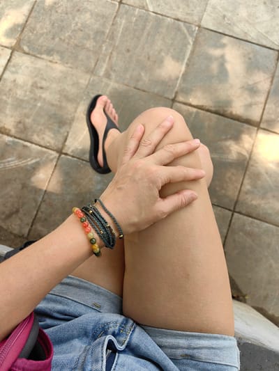 A person is seated outdoors on a tiled surface. The individual is wearing denim shorts, a pink bag, and black flip-flops. They have bracelets on their left wrist. Their left hand rests on their right knee. The location is indicated as Thành phố Hà Giang, Vietnam.