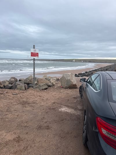 The image presents a coastal scene at Montrose Beach, Montrose, United Kingdom, under an overcast sky. In the immediate foreground on the right, the rear passenger side of a dark-colored two-door coupe vehicle is partially visible, parked on a reddish-brown dirt and sandy surface exhibiting tire tracks. To the left of the vehicle, a formation of large, irregular rocks acts as a natural barrier, with a notably large, light-colored boulder positioned centrally among them. A grey metal pole rises from this rocky area, supporting a rectangular white sign with a red border. The sign displays a red 'no dogs' pictogram and the prominent text "DOG CONTROL," followed by smaller, partially legible regulatory text.

Beyond the rocks and sign, the North Sea is visible with breaking waves approaching a sandy beach. On the distant horizon to the left, a tall, white lighthouse (North Esk Lighthouse) stands. Further along the coast to the right, a smaller white lighthouse, accompanied by white buildings (Montrose Lighthouse), marks the entrance to a body of water, likely the Montrose Basin or River South Esk. The distant coastline consists of low-lying, green-brown hills. The sky is uniformly grey with heavy cloud cover. No individuals are present in the scene.