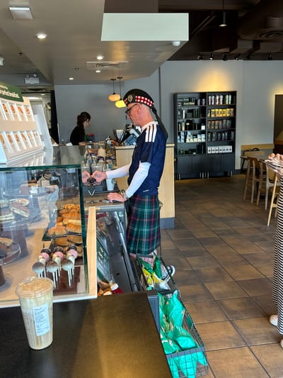 The image depicts the interior of a Starbucks in Pitt Meadows, Canada. A man wearing a kilt and a tartan hat is standing behind the counter, holding what appears to be a payment device and looking towards a customer. The customer's hands are visible, reaching for a drink or food item.

In the foreground, a glass display case showcases various pastries and desserts, including croissants, cookies, and cake pops. To the left, a cup of iced coffee with a Starbucks label sits on the counter. Behind the counter, a woman wearing a black shirt is visible, potentially a barista. Further back, shelves are stocked with Starbucks merchandise, including coffee beans and tumblers. The flooring is tiled, and tables and chairs are set up in the background, suggesting a seating area. A wire basket filled with green bags of snacks is also visible near the counter. The overall lighting suggests it is daytime.