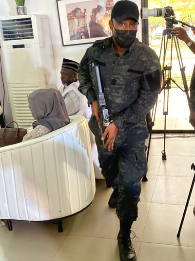 A man wearing a black camouflage uniform, a black mask, and a black cap is lying on the floor. He is holding a silver and black rifle. Behind him are two men sitting on a white tufted sofa. One of the men is wearing a white shirt and a black and gold cap. The other man is wearing a gray hoodie. Behind them is an air conditioner, a framed picture, and a white sofa. In the background is a camera on a tripod. The photograph was taken in Kwarau, Nigeria.