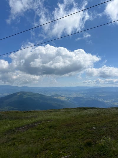The image depicts a panoramic view of mountainous terrain under a bright blue sky filled with fluffy white clouds. In the foreground, a grassy slope leads down towards a vast expanse of layered, dark blue mountain ranges stretching to the horizon. The mountains are covered in dense vegetation, with some areas appearing as shadows. The sky is dominant in the upper portion of the image, with three dark, parallel lines running diagonally across it, suggestive of power lines or a ski lift cable. The scene appears to be a natural, undeveloped landscape. There are no people or animals visible. The lighting suggests it is daytime, likely late morning or early afternoon.