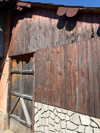 The image shows the exterior of a building in Valchedram, Bulgaria. The wall is composed of weathered, horizontal wooden planks, except for a section on the right side which is covered in light-colored, irregularly shaped stones. There is a small, wooden shuttered window near the bottom. The roofline is partially visible with decorative red tiles along the edge. A clear blue sky can be seen in the upper left corner.