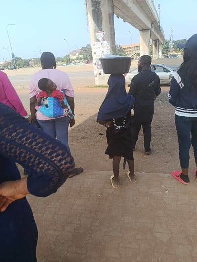 The scene depicts a street-level view in Gbessa, Nigeria, beneath an elevated concrete overpass. Five individuals are visible, primarily from behind.
In the immediate foreground, a person's left arm and torso are partially visible, wearing a dark blue garment with a textured, possibly lace, pattern.
Further back, from left to right:
1. An adult female, seen from the back, wears a light pink t-shirt and blue jeans. She carries a baby on her back in a blue and pink patterned baby carrier. The baby wears a red top and its head is turned to the right.
2. A child, also seen from the back, wears a dark blue hijab covering her head and a dark dress. She is balancing a large, dark-colored basin on her head. She wears light-colored sandals.
3. An adult male, seen from the back, wears a black long-sleeved shirt and black trousers. A black bag is slung across his shoulder. He holds a small, clear bottle in his right hand.
4. Partially visible on the far right, an individual wears a blue cap, a blue and white striped jacket, and blue jeans. Pink slides are on their feet.
The ground consists of paved sections in the foreground transitioning to dirt/gravel beneath the overpass. A large concrete pillar of the elevated structure stands in the midground, covered with several posters or fliers. In the background, a multi-lane road is visible with several vehicles (cars) and distant buildings and trees under a clear, bright sky.