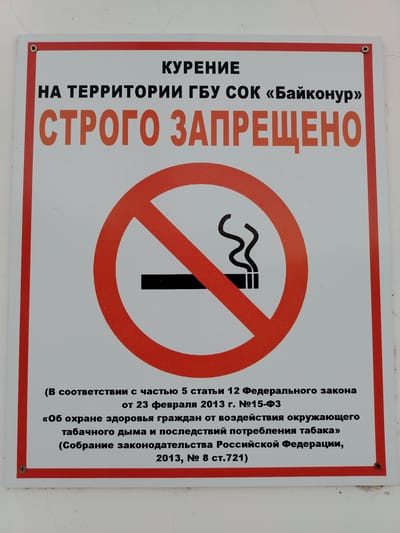 The image shows a "No Smoking" sign in Russian. The sign is red and white and features a graphic of a cigarette with smoke, crossed out by a red circle and diagonal line. The text at the top reads " курение на территории ГБУ СОК «Байконур» строго запрещено" which translates to "Smoking on the territory of the State Budgetary Institution of Social Services "Baikonur" is strictly prohibited." Below the graphic, there is smaller text in Russian that cites legal regulations related to the prohibition of smoking. The sign is mounted on a white wall, likely a building exterior, in a public area. The lighting suggests it is daytime. There are no people or other significant objects visible in the frame, other than the sign itself and the wall it's attached to. The context of "Baikonur" in the text indicates the location is the Baikonur Cosmodrome, which is a leased Russian launch facility in the Kyzylorda Region of Kazakhstan.