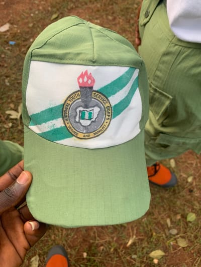 A light olive-green baseball cap, featuring a white front panel with diagonal green stripes and a circular emblem, is held by a dark-skinned hand in the lower-left quadrant. The emblem displays a flaming torch above a map of Nigeria, encircled by the text "NATIONAL YOUTH SERVICE CORPS" and "SERVICE AND HUMILITY." In the background, a person wearing light olive-green uniform trousers and a white undershirt is partially visible on the right, with their left foot, shod in an orange shoe with a grey toe, visible on the ground. The environment consists of reddish-brown soil sparsely covered with dry green and brown vegetation. The specific location within Kubi A, Nigeria, is not discernable from the visual information presented.
