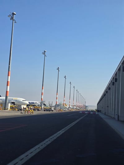 The image depicts an airport apron at Berlin Brandenburg Airport (BER) in Schönefeld, Germany, under a clear blue sky. A linear arrangement of approximately fifteen tall metal poles with red and white striped lower sections extends into the background from the left midground, each topped with a spherical light fixture and multiple directional lights/antennae. To the left, two commercial passenger aircraft are parked. The front fuselage and nose section of a white Airbus A321 aircraft, bearing "eeling.com" on its livery, is prominent, with mobile passenger stairs positioned at its forward entry. Various yellow and black ground support equipment, including baggage carts and safety barriers, are staged around it. A second, partially visible white aircraft with "AIR" visible on its fuselage is positioned behind the first. To the right, a large, modern airport terminal building with a grey, vertically segmented facade runs parallel to the tarmac, extending into the background. A concrete walkway borders the terminal building, where a group of several individuals, clad in dark clothing, are visible near the distant end. The foreground comprises dark asphalt pavement with a linear drainage grate and white painted markings. Further down the tarmac, a series of white and pink plastic barriers are positioned. The scene indicates active airport operations with parked aircraft and ground infrastructure in place.
