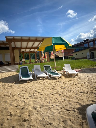The image depicts a sunny outdoor scene with a sandy beach in the foreground. Several white plastic lounge chairs are arranged on the sand, some with green fabric seats. A colorful yellow and green striped umbrella provides shade over a portion of the beach area, with other patterned umbrellas visible underneath. In the background, a building with multiple windows and a green lawn can be seen. The sky is a vibrant blue with scattered white clouds. The image appears to be taken during the daytime on a clear day. The setting suggests a recreational area, possibly a resort or a public beach. There is no activity or event taking place, and no people are visible. The overall mood is relaxed and inviting, typical of a summer day at the beach.