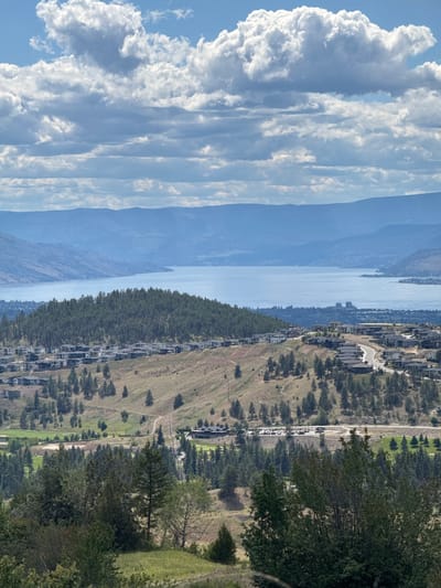 The image depicts a panoramic view of a developed hillside overlooking a large body of water, likely a lake, with mountainous terrain in the distance. The foreground features rolling hills with scattered evergreen trees and patches of dry grass. A residential development of modern-style houses with dark roofs is visible cascading down the hillside. Further down the slope, a golf course or park area with green fairways and trees is present, along with a road and a parking lot filled with vehicles. In the midground, the expansive blue water of the lake stretches towards the horizon, bordered by more distant hills. The sky is partly cloudy, with large, puffy white clouds set against a bright blue sky. The overall scene suggests a developed suburban or exurban area integrated with natural landscape, characteristic of the East Central Okanagan Electoral Area in Canada. No people are clearly visible in the image.