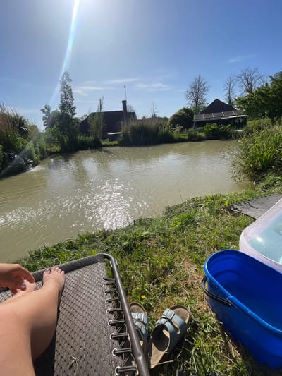 This image captures a serene outdoor scene on a bright, sunny day, likely beside a canal or river in Ashford, United Kingdom. The perspective is from a reclining position, looking across the water.

In the foreground, on the right side of the frame, a person is relaxing with their bare legs and feet propped up on a dark-patterned lounge chair. A hand is also visible near their knee. Immediately to the right of the chair, on the grassy bank, are a pair of light blue buckled sandals, a deep blue bucket with a handle, and a lighter, semi-transparent plastic tub or basin. The grass in this area is green with some drier patches, and a small yellow flower is visible.

The middle ground is dominated by a calm, murky brown body of water, stretching diagonally across the frame. The surface of the water reflects the intense sunlight, creating numerous sparkling highlights and ripples. The opposite bank is densely lined with various green vegetation, including reeds and trees, some of which appear deciduous.

In the background, beyond the foliage on the far bank, several residential-style buildings with dark roofs are visible. One building features an antenna or mast extending upwards. The sky above is a clear, vibrant blue, indicative of excellent weather. A prominent sunflare with rainbow prismatic effects is visible in the upper left quadrant of the sky, emphasizing the brightness of the day.

The overall scene conveys a sense of peaceful relaxation and enjoyment of warm, sunny weather by the water. There is no visible text in the image.
