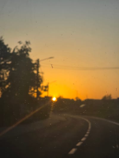 The image shows a view through a dirty window, possibly of a vehicle. The scene outside depicts a road with white lines on the edge, suggesting it could be in a residential area or on the outskirts of a city. There are trees visible, silhouetted against a bright orange sunset or sunrise. The light suggests the photograph was taken during twilight. There are no people visible in the image. The city of Mouscron, Belgium is not apparent from the content of the image.