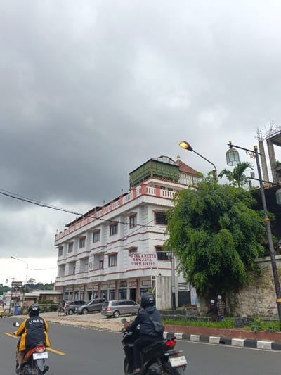 The image captures a street view in Palayan City, Philippines, under an overcast sky. Dominating the center is the "HOTEL & RESTO SENJAYA," a multi-story building painted white with pinkish-red accents around its windows and balconies. A large green structure and a small domed feature are visible on its roof. A sign affixed to the building reads "HOTEL & RESTO SENJAYA (0263) 519797."
In the immediate foreground, two individuals on motorcycles travel away from the viewer on an asphalt road. The motorcyclist on the left wears a black helmet and a yellow and black jacket with "UBER" emblazoned on the back; their motorcycle's license plate is "9023-TE." The motorcyclist on the right wears a black helmet and dark attire; their motorcycle shows part of license plate "F 6410 ND." The road is marked with a solid yellow line.
To the right of the road, a large, dense green tree is prominent, partially obscuring a stone wall. Behind this wall and the tree, two figures are partially visible, both wearing white head coverings and patterned brown garments. An illuminated streetlamp with a yellow glow and a decorative lantern-style streetlamp are positioned on the right side of the image. Several vehicles are parked on the ground level in front of the hotel, and overhead power lines traverse the upper portion of the scene.