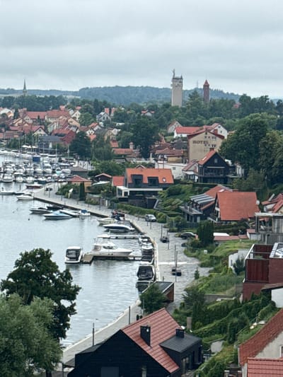 The image depicts a panoramic view of the town of Mikołajki, Poland, showcasing a harbor filled with numerous docked boats. The scene is characterized by a dense arrangement of buildings with red-tiled roofs, typical of European coastal towns. In the background, a distinctive tower, possibly a water tower or observation deck, stands tall amidst lush greenery and a cluster of trees. The water in the harbor is calm, reflecting the overcast sky, which suggests a moderate weather condition. A paved walkway runs along the harbor's edge, providing access to the boats. Some of the buildings appear to be residential houses, while others might be commercial establishments or hotels, as suggested by the visible "HOTEL" sign on one building. The overall impression is one of a well-established lakeside community with a strong connection to water-based activities.