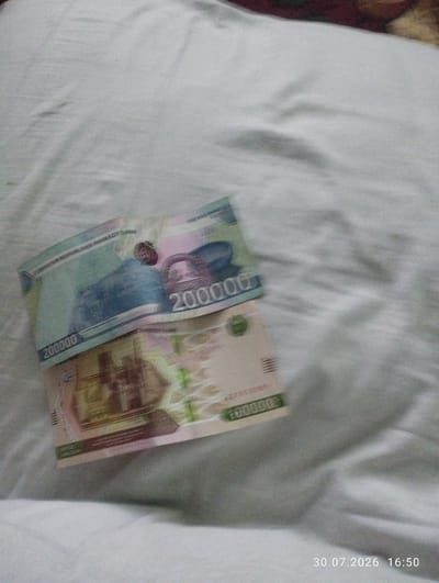 The image shows two banknotes, which appear to be from Uzbekistan, lying on a white, rumpled surface that resembles bedding. The banknotes are partially overlapping and angled. The top banknote is a light blue and green hue, featuring a large number "200000" and text that seems to read "O'ZBEKISTON RESPUBLIKASI MARKAZIY BANKI". The second banknote, partially visible beneath the first, is a lighter color with pink and green accents and also displays denominations, including "200000" and what appears to be a building or monument. The lighting is dim, and the image has a slight blurriness, suggesting it was taken indoors. A timestamp in the lower right corner indicates "30.07.2026 16:50". There is no indication of a city or specific location within Quyosh, Uzbekistan.