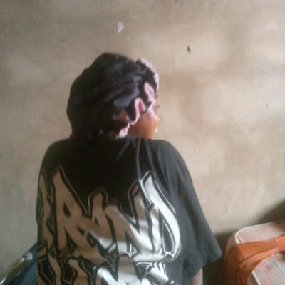 The scene depicts an indoor setting in the city of Ibreda, Nigeria. A single individual is positioned with their back partially to the viewer, facing towards the right side of the frame. The person is wearing a dark, possibly black, satin head covering with a visible light pink or beige ruffled edge. A black short-sleeved t-shirt is worn, featuring a large white graphic design on the back composed of stylized, thick lettering with a dark outline. The background is a plain, light brown or beige wall with some minor marks. In the lower right corner, a portion of orange-brown upholstered furniture is visible.