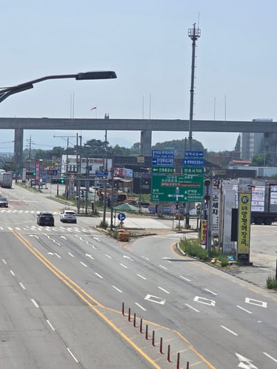 The image depicts an overhead view of a moderately busy street intersection in Pyeongtaek-si, South Korea, under a clear, bright sky. A multi-lane road curves to the right, with vehicles including cars and trucks navigating the intersection. Several traffic lights are visible, some displaying green signals. Numerous road signs, predominantly in Korean, provide directional information to various locations and services, including "Free Trade Zone," "Pyeongtaek Port," and "Pyeongtaek Lake Resort." Along the roadside, there is sparse vegetation and several utility poles. In the background, a large overpass or elevated highway structure dominates the skyline, with a communication tower positioned further in the distance. The overall scene suggests an urban or semi-urban environment with ongoing traffic flow.