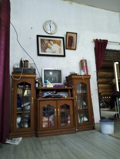 The image depicts an interior scene of a room, likely a living room or entertainment area, in Aluedua, Indonesia. The walls are painted white and appear textured. A large wooden cabinet dominates the center of the frame, featuring glass-fronted doors on both the upper and lower sections. The upper part of the cabinet holds a vintage television set, an iron, and some other electronics. The lower cabinets display various items behind the glass, including what appear to be bowls and fabrics.
To the left of the cabinet, a dark purple curtain hangs. Above the cabinet, a round analog clock with Roman numerals is mounted on the wall, along with a framed photograph of two individuals, and another framed artwork. To the right of the cabinet, a tall, red plastic container with a floral pattern stands on a smaller wooden unit. Beyond this, a doorway leads to another part of the house, showing wooden walls and horizontal wooden planks, suggesting a rustic construction style. A bicycle is partially visible in the background. On the floor to the far right, a white and blue plastic container is present. The floor itself has a light-colored, marbled or striped pattern. The overall impression is that of a lived-in, modestly furnished home.