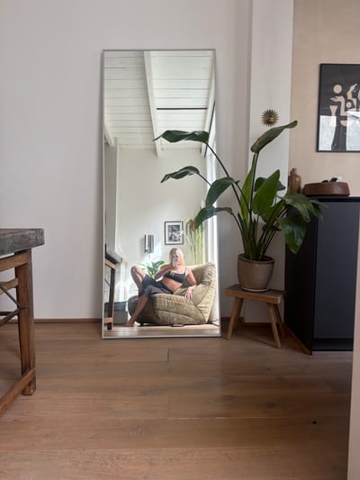 A full-length mirror stands upright against a white wall in what appears to be a residential interior. In the reflection of the mirror, a person with blonde hair is seated in a textured, olive-green armchair, holding a phone and taking a selfie. The person is wearing a black bralette and grey patterned shorts. The mirror also reflects a wooden floor, a white slatted ceiling, and a framed piece of abstract art on a beige wall. To the right of the mirror, a tall potted plant with large green leaves sits on a small wooden stool. Behind the plant, a dark cabinet is visible, with a wooden bowl and a ceramic vase on top. To the left of the mirror, a portion of a rustic wooden table with a woven seat is visible. The overall impression is one of a modern, casually decorated living space. The image does not contain any information that would suggest a specific location in Leiden, Netherlands.