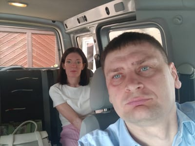 The image shows two people, a man and a woman, in what appears to be the interior of a van or minibus. The man is in the foreground, taking a selfie with a blue collared shirt. He has light blue eyes and short brown hair. The woman is seated behind him to his left, wearing a white t-shirt and pink striped pants. She has dark brown hair. The interior of the vehicle shows gray seats and a white ceiling light. Outside the vehicle, through the windows, wooden siding and a building are visible. The exact location within Altdorf bei Nürnberg, Germany, is not discernible from the image.