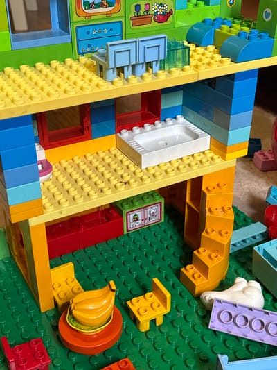 The media file displays a close-up, overhead view of a collection of brightly colored plastic toy bricks, primarily in shades of yellow, blue, red, and green, arranged on a green baseplate. The bricks are consistent with the style of building blocks designed for young children.

Several distinct objects are constructed or represented using these bricks. Prominently featured is a white toilet with a pink lid, positioned on a yellow surface. Adjacent to this is a yellow structure with steps, and further to the right, a cluster of bright yellow banana-shaped bricks sits within an orange circular piece. Other visible elements include a white, cloud-like plastic shape, a purple rectangular brick with circular indentations, and a green and white sticker depicting a cat wearing a party hat and holding balloons. There are also several smaller, single-color bricks and some translucent blue pieces.

The arrangement suggests a partially built structure or a scene created from these toys. The background is a green baseplate with the characteristic circular studs used for connecting toy bricks. The overall impression is one of a children's play area, with the toys meticulously arranged on a play mat or floor. There are no discernible people, animals, or indications of time of day or weather. The location context provided is Zassovskaya, Russia, but this cannot be visually confirmed from the image itself. There is no visible text within the image.