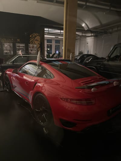 The image shows a red Porsche, possibly a vintage model, parked inside a building in Wetzikon, Switzerland. In the background, there is a wall decorated with a lion emblem and framed vintage automotive photographs. The building's architecture features high ceilings and visible structural beams. Another car is also parked nearby.