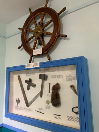 The image shows a museum display case with various nautical and woodworking tools, likely related to boat construction. Above the case, a large, ornate wooden ship's wheel is mounted on a light blue wall. A white placard is attached to the wall below the wheel. The display case itself has a blue frame and contains several items, including a hammer, a wooden mallet, a bundle of dark, coarse hair, a piece of wood, and several metal and wooden implements. Some of the tools are labeled with small white placards in Spanish. One of the placards indicates the donation was made by the family of Captain Rodríguez Cedeño in memory of Captain Francisco Rodríguez, owner of the ship Carmen Felicia, and that the donation was made in Boca del Río on March 19, 2016. The presence of the ship's wheel and the context of nautical tools suggest a maritime museum or historical exhibit. The overall environment appears to be indoors, with neutral-colored walls.
