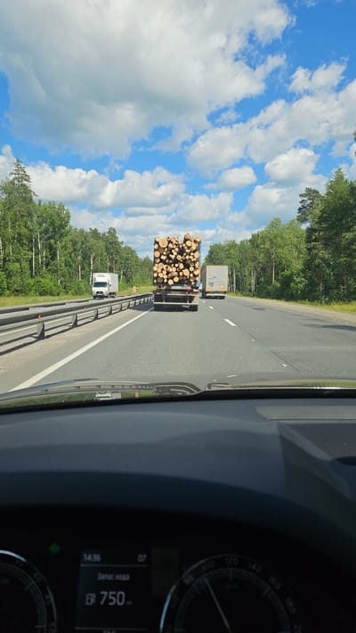 This image shows a view from inside a vehicle, looking out at a highway in Voskresenskoe, Russia. The scene depicts two trucks driving on the road, one of which is carrying a large load of cut logs. The sky is a vibrant blue with scattered white clouds, indicating fair weather. Lush green trees line the sides of the highway, suggesting a rural or semi-rural setting.

The perspective from the driver's seat includes a portion of the car's dashboard on the right side of the image. A speedometer with markings up to 240 is visible, and a digital display shows "14:36" and "750". Another, smaller text reads "Запас хода". The time of day appears to be mid-afternoon. The overall impression is that of a daytime journey on a highway through a forested area. There are no people clearly visible.