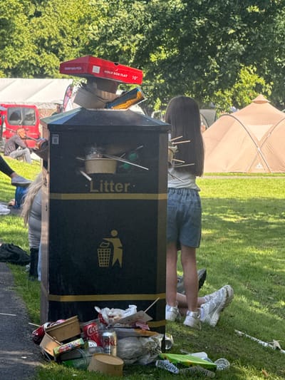 The image captures an overflowing public waste bin with the word "Litter" prominently displayed on its side. The bin is situated outdoors in a grassy area, likely a park or public space, with trees and tents visible in the background, suggesting an event or festival. A young girl with dark hair, wearing denim shorts and a white t-shirt, stands near the bin, her back to the camera. Around the bin, there is a significant amount of scattered refuse, including cardboard pizza boxes, drink cans (Coca-Cola and a silver can labeled "This is white"), plastic bottles, and various food packaging. In the middle ground, people are seen sitting on the grass, and a red vehicle is parked to the left. The overall scene depicts a public area suffering from littering and overflowing waste facilities.