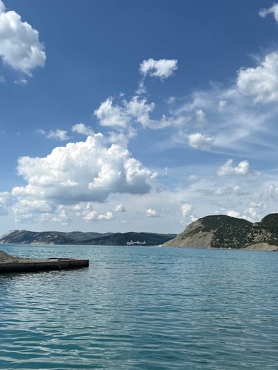The image presents a detailed view of a coastal environment in Bol'shoi Utrish, Russia, under a clear, partially clouded sky. The upper two-thirds of the frame are filled with a vibrant blue sky, populated by numerous white cumulus clouds varying in size and density, with a prominent, large cloud formation occupying the upper-mid left quadrant.

The lower third of the image is dominated by an expanse of calm, teal-blue water, likely the Black Sea, displaying gentle ripples and reflections across its surface. In the lower-left foreground, a dark, flat, rectangular structure, consistent with a weathered concrete or stone pier, protrudes partially into the water. Its visible surface shows signs of wear and is topped with some darker, granular material where it meets the unseen shoreline.

A chain of undulating mountains or large hills forms the backdrop, extending across the horizon. The slopes exhibit varying textures and hues, with some sections near the water appearing lighter in color, possibly due to rock or sparse vegetation, while other areas are covered in denser, dark green foliage. Along the distant mountain ridges, several faint, white, slender structures, consistent with wind turbines or communication towers, are discernible. On the far right, at the base of the mountains along the coastline, a cluster of white buildings suggests a small settlement or resort area. No human figures are visible.