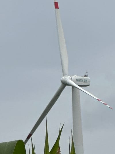 The image is a close-up, low-angle shot of a wind turbine against a cloudy, overcast sky. The wind turbine is predominantly white with some red accents on the blades and a red band at the top of the tower. The word "SUZLON" is visible in blue lettering on the nacelle of the turbine. The blades of the turbine are white with red stripes near their tips. In the foreground, there are green plants that appear to be corn stalks, obscuring the base of the turbine. The scene suggests an agricultural or open landscape setting. There are no people or animals visible in the image. The image was taken in Kolubaivtsi, Ukraine.