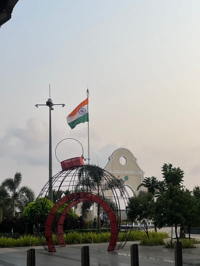 The image shows the Indian flag waving atop a tall flagpole in Kasarvarne, India. A surveillance camera is mounted below the flag. To the right, there is a large, abstract metal sculpture. In the background, there's a light-colored building with an arched entrance. Various trees and greenery surround the scene. The sky is overcast and gray.