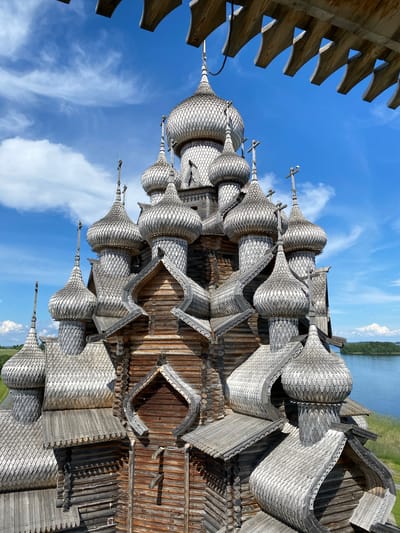 The image shows a large, ornate wooden church with multiple domes and intricate carvings. The church is made of logs and has a gray, weathered appearance. The domes are covered in small, overlapping wooden shingles and are topped with crosses. The church is situated in a scenic location with a body of water and trees in the background. The sky is blue with scattered white clouds. The architecture appears to be traditional Russian Orthodox. The image is taken from an elevated perspective, looking down slightly at the church, and a wooden overhang is visible in the foreground. The location is identified as Boyarshchina, Russia, likely referring to Kizhi Island, famous for its wooden architecture. There are no people visible in the image.
