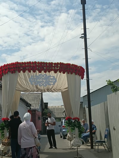 In Osh, Kyrgyzstan, a wedding arch adorned with red flowers and white drapery stands over a street. "Wedding Day" is inscribed on a white cutout attached to the arch. Several people are present. A man in a white t-shirt and black pants holds a camera on a tripod, appearing to film. Two older men in caps sit on chairs near a corrugated metal fence. A woman in a pink hijab and a patterned skirt stands with her back to the camera. Other individuals are also visible, including a child. Buildings with corrugated roofs line the background. Overhead, power lines crisscross against a cloudy sky. A utility pole with several horizontal attachments stands to the right of the frame. A car is parked in the background.
