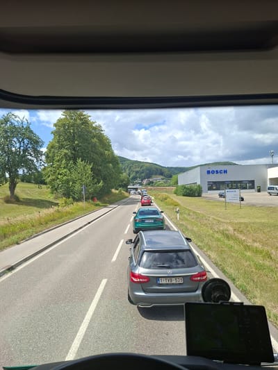 The image, taken from inside a vehicle, shows a traffic jam on a road in Bad Ditzenbach, Germany. In the foreground, a dark gray Mercedes-Benz station wagon with license plate 1-TVB-539 is stopped. A black GPS device is mounted on the dashboard in front of it. Ahead, several other cars are also stopped in traffic. To the right of the road is a grassy field with a modern building in the distance featuring a large blue "BOSCH" logo. To the left of the road, there is a grassy embankment and trees. The sky is partly cloudy.