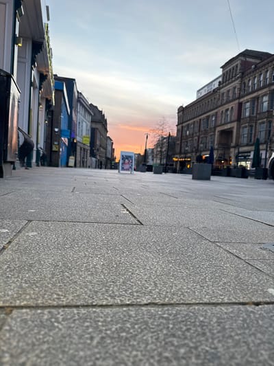 This low-angle photograph captures a pedestrianized section of Fishergate in Preston, United Kingdom, at sunset. The foreground is dominated by speckled grey paving slabs with visible grout lines, leading into the distance.
On the left, a row of commercial buildings features varied shopfronts; one in the immediate foreground has a white facade and partial illumination. Further down, blue and grey building facades are discernible. A black street bin is partially visible on the far left. Two indistinct human figures are present near the left-side buildings; one appears to be bent over.
On the right, a multi-story historic building with a prominent brick and stone facade, ornate architectural details, and arched ground-level entrances dominates the mid-ground. Dark outdoor dining umbrellas are visible outside establishments on this side, interspersed with square, dark street furniture, possibly planters or bollards.
Centrally, an illuminated A-board or display stand with colorful graphics stands in the mid-distance. The street recedes into the background, where bare tree branches and distant streetlights are silhouetted against the horizon. The sky, which occupies the upper third of the image, transitions from a pale blue directly overhead to vibrant orange and pink hues at the western horizon, indicating the time is dusk. The scene is largely devoid of significant human activity or interaction.