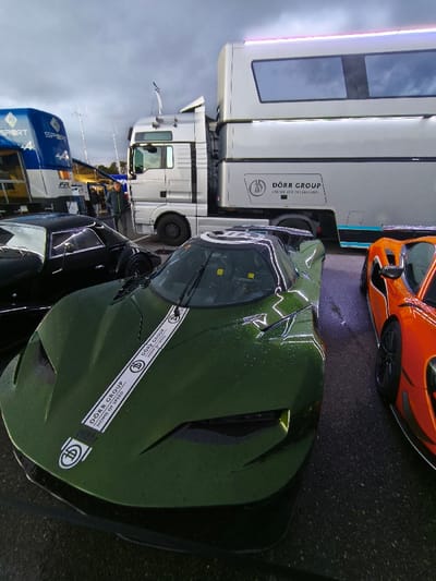 This image depicts an outdoor vehicle staging area, likely a paddock at the Hockenheimring racetrack in Hockenheim, Germany, under an overcast sky with a wet asphalt surface.

In the immediate foreground, a dark green, low-slung, high-performance automobile with aerodynamic styling is positioned centrally, facing forward. Its hood bears a longitudinal white stripe with "DÖRR GROUP HOME OF SPEED" branding. The vehicle's surface is covered with numerous small water droplets. To the right of the green vehicle, the front-right section of a bright orange, similarly aerodynamic sports car is visible, also exhibiting water droplets. To the left and slightly behind the green vehicle, the rear-left quarter of a glossy black, coupe-style sports car is partially visible.

In the mid-ground, a silver MAN semi-truck cab is coupled to a large, multi-level silver transport trailer. The trailer features "DÖRR GROUP PORSCHE FULDA RHEINMAIN" branding, reflective windows on its upper section, and blue and black trim along its lower rear. Between the black sports car and the semi-truck, a male individual in a dark jacket and light trousers stands facing towards the right side of the frame. In the left background, a portion of a blue and white transport vehicle or trailer with "RENAULT SPORT" and "ALPINE" branding is visible. The scene suggests a motorsport or automotive event occurring in damp conditions.