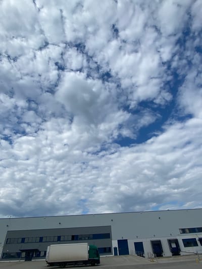 The image shows a large, modern industrial warehouse building located in Bielany Wrocławskie, Poland. The building has a light gray facade with dark blue window frames and multiple loading docks. In the foreground, a white box truck with a green cab is parked on the asphalt in front of the warehouse. The sky above is filled with a dramatic arrangement of white and gray clouds against a bright blue sky. The scene appears to be a commercial or industrial area under a partially cloudy sky.