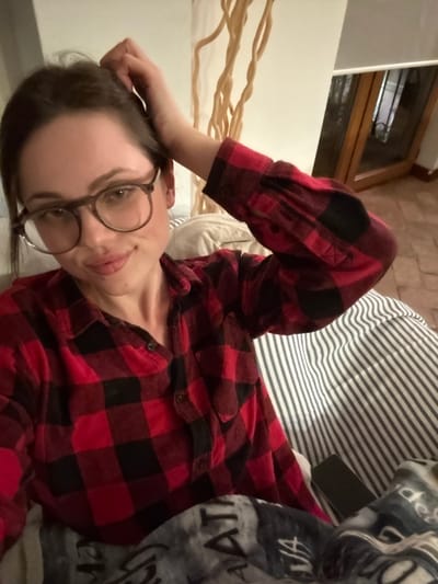 An indoor scene in Cava de' Tirreni, Italy, depicts a Caucasian female subject positioned centrally. She has dark hair, wears eyeglasses with dark frames, and a red and black plaid flannel shirt with the sleeves partially rolled up, revealing black elbow patches. Her left hand is raised, fingers lightly touching the hair near her left temple. Her right arm is bent, resting on a surface covered by a white and dark grey striped fabric, likely a pillow or bedding. A dark grey/black blanket with white text (partially visible as "AT" and "VIA") covers her lower body. A black mobile phone is visible resting on the striped fabric to her right. The subject exhibits a slight smile and looks directly forward. The background features a light-colored wall with a tall, light-colored decorative element composed of intertwining, branch-like structures. To the right, a wooden-framed window or door with glass panes is partially visible, topped by a white roller blind. A reflection in the glass suggests a tiled floor with reddish-brown hues. The lighting is soft and ambient.