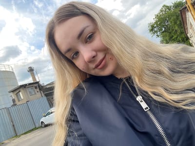 A woman with long blonde hair is wearing a black bomber jacket. She is smiling at the camera. In the background, there is a metal fence, a white car, a small building with a window, and a taller structure with a chimney. A tree with green leaves and part of a building are also visible. Based on the buildings and objects present in the image, it appears to be taken in an industrial area within the city of Bobrov, Russia.