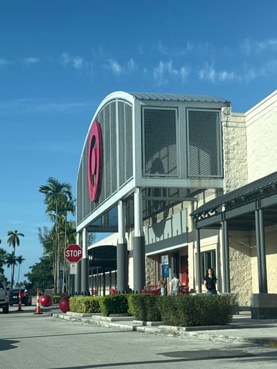 The image captures the exterior of the Target retail store located at 21205 Biscayne Boulevard in Aventura, Florida, United States, observed from a paved parking lot or access road. The scene is set under a clear blue sky with sparse, wispy white clouds, indicating daylight conditions.
The prominent structure is the store's main entrance, featuring an elevated, arched facade with a large, red, circular Target bullseye logo. This arched section is constructed with a metal mesh screen and topped by a corrugated metal roof, supported by four dark gray cylindrical pillars. The building's walls consist of light-colored blockwork and stucco sections. To the right of the main entrance, a darker-colored canopy extends over a pedestrian area, supported by black rectangular columns.
In the foreground, a paved surface leads towards the building, bordered by concrete curbs and low planter beds containing green, manicured bushes. A red octagonal STOP sign on a pole is positioned to the left of the main entrance. On the paved surface, two large, red, spherical objects, possibly decorative bollards, are situated near the planter bed, with an orange traffic cone placed between them. The front portion of a light-colored SUV or truck is partially visible on the far left.
Three individuals are visible near the store's entrance. One person, wearing a dark top, stands on the right side of the main entrance. Two other individuals, one in a light shirt and shorts and the other in a light top and shorts, are closer to the entrance, near what appears to be red outdoor seating, with their backs partially or fully to the viewer. Several tall palm trees are visible on the left side of the frame, behind the STOP sign. Shadows cast by the building and objects are visible on the ground, indicating the sun's position.