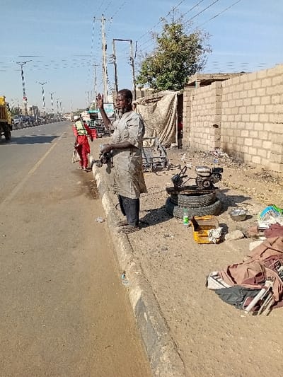 The image depicts a daytime outdoor scene in Sharada, Nigeria, alongside a paved multi-lane road under a clear blue sky. An adult male, of apparent African descent, stands on an unpaved shoulder bordering the road in the foreground. He wears a heavily soiled light-colored tunic over dark trousers and sandals. His right hand is raised with an open palm, and his left hand holds a dark, elongated object.
Adjacent to him on the ground, mechanical components are visible: a small engine mounted on a frame rests on two dark-colored tires. Nearby are a yellow plastic crate, a green plastic bottle, a metal bowl, and assorted piles of fabric and debris. To the right of the scene, a partially constructed light-colored block wall and a makeshift structure covered with a light-colored tarp are situated. A tree with green foliage is visible behind these structures. Overhead utility lines and poles run parallel to the road.
In the mid-ground on the paved road, a second individual, seen from behind, walks away from the viewer. This person is dressed in red clothing, overlaid with a yellow reflective vest or harness, and appears to carry a light-colored bag or object in their left hand. On the far left of the road, a yellow dump truck is partially visible, moving away from the camera. Additional vehicles and modern streetlights are discernible in the distant background, indicating an urban or semi-urban roadway.