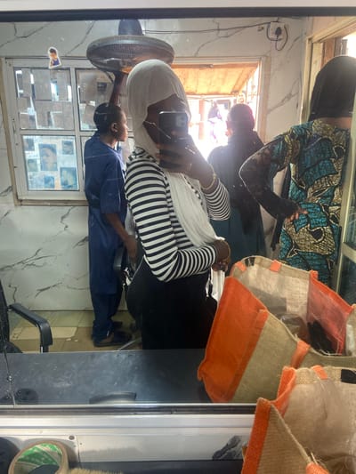 A reflection in a mirror shows the interior of a hair salon in Okengwe-Obehira, Nigeria. A woman wearing a black and white striped top and a white head covering is taking a photo with her cell phone. A person in a blue uniform is working on the hair of a seated customer. Another person with a black head covering and a patterned turquoise and black dress is standing nearby. A fan is visible on the left. The background includes a window covered with photos. Orange-trimmed burlap bags hang at the top right.